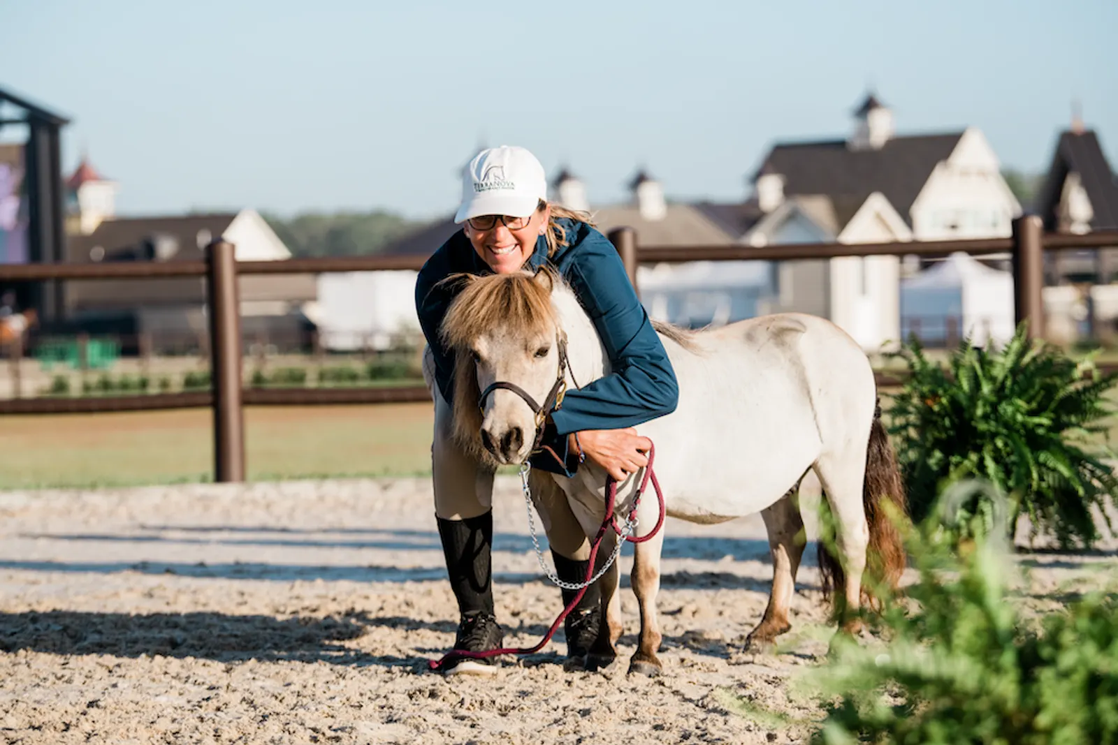 A smiling handler in a TerraNova cap hugging Fabio, a white miniature therapy horse, in the arena at TerraNova Equestrian Center. Photo by EQ Media.