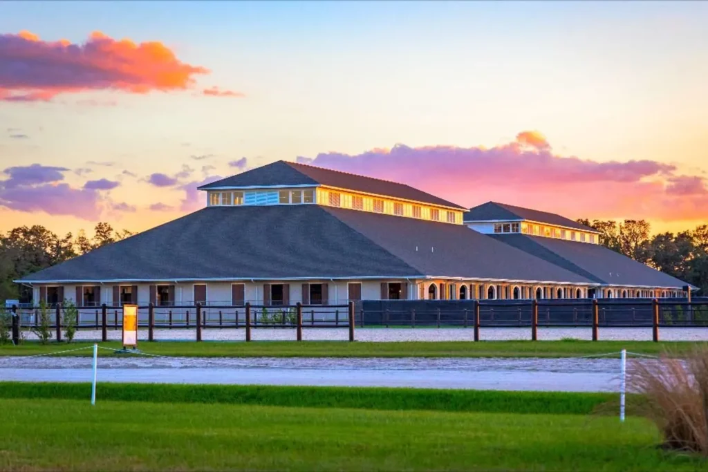 Permanent stabling barns at TerraNova Equestrian Center glowing at sunset behind dark wood fencing and a groomed arena